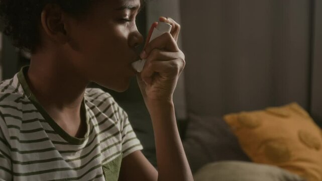 Close up view of young Black boy using inhaler device to prevent asthma attack while sitting at home