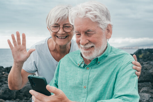 Portrait of smiling bonding senior couple sitting outdoors on the seaside video calling by smartphone. Cloudy sky in background and horizon over water