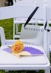 Open lilac fan and yellow rose lie on white chair outside. Close-up of lilac fan made of light wood. Chinese folding fan for cooling in hot weather without electricity. Reserve sign on chair