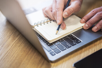 A man's hand writing in a notepad on a laptop, captured in close-up detail, set against a blurred surrounding