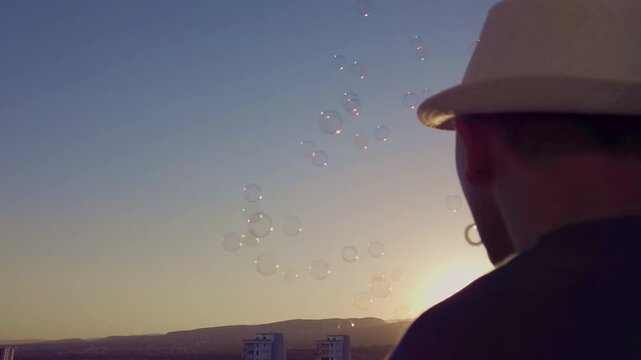 Silhouette of a man in a white hat against a sunset sky, blowing soap bubbles, rear view, camera movement