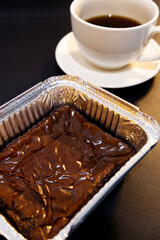 Brownies in foil cups with blurred coffee cup placed on black background. Close-up chocolate brownie cup and hot coffee in vertical. Homemade, dessert at home, snack time, and warm light concepts.