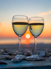 Two glasses of wine at sunset create a romantic scene with oyster shells scattered around, evoking relaxation.