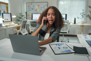 Obraz premium Confident African American businesswoman talking on the phone in the office. Professional young woman contacting, discussing business, investment plan, working on laptop, communication concept.