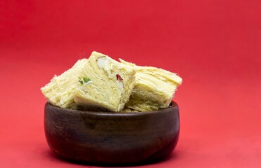 Soan Papdi or Patisa Garnished with Cashew Nut, Pistachio, Almond and Saffron in Wooden Bowl Isolate on Red Background with Copy Space, Also Known as Shompapri or Shonpapdi
