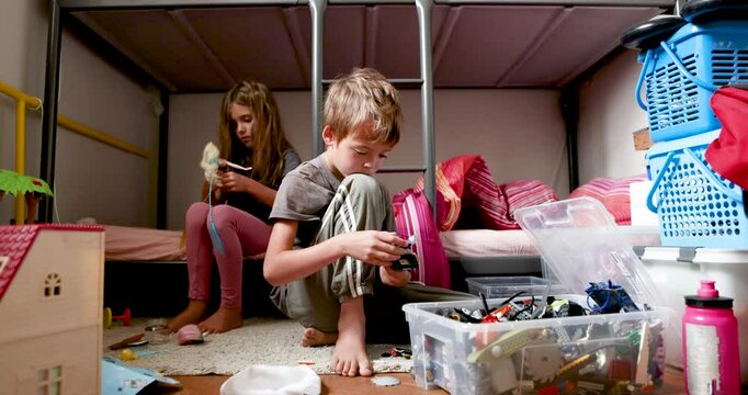 Real time of concentrated girl looking down while sitting on bunk bed adjusting dress on small doll with hands in daylight and little brother sitting near on floor playing with toys in box small room