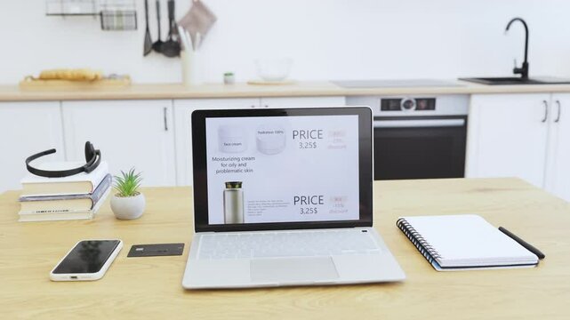 Open laptop showing website of cosmetics products with discount prices in home setting. Features moisturizing cream for oily and problematic skin. Modern workspace with notepad, smartphone, and decor.
