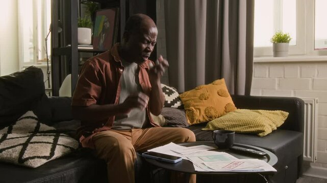 Stressed Black man sitting on sofa in living room, reviewing overdue bills, coughing and using asthma inhaler