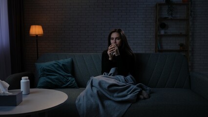 Fototapeta premium Woman suffering mental health issues, sitting on the sofa covered in blanket, depressed expression, calming down, drinking glass of water.
