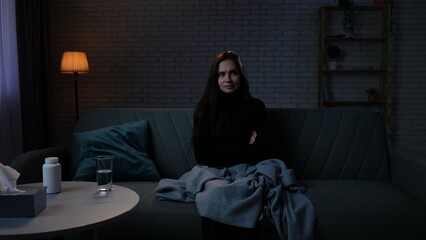 Fototapeta premium Woman suffering mental health issues, sitting on the sofa covered in blanket, depressed expression, looks at side suspicious face, closed hands gesture.
