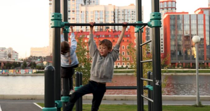Tracking shot siblings conversing while playing in calisthenics park and hanging on parallel bars and then boy climbing down to pick scooter during evening time against city buildings river