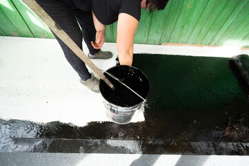 A woman uses a brush to apply bitumen to a concrete floor.