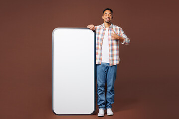 Full body young happy man of African American ethnicity he wear blue shirt casual clothes big huge blank screen mobile cell phone smartphone with area show thumb up isolated on plain brown background.