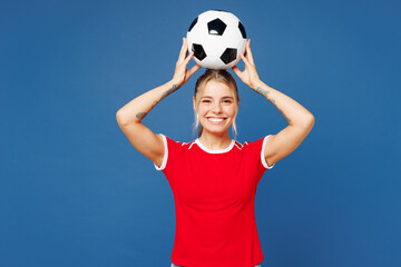 Young smiling cheerful happy woman fan wear basic red t-shirt cheer up support football sport team hold in hand soccer ball watch on head tv live stream look camera isolated on plain blue background.