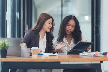 Collaborative Businesswomen Reviewing Project: Two diverse businesswomen, one Asian and one African-American, collaborate intensely over a tablet in a modern office setting.