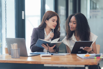 Collaborative Businesswomen: Two diverse businesswomen engage in focused discussion, reviewing documents and using digital devices in a modern, well-lit office setting.