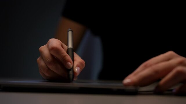 Graphic designer drawing on tablet with digital pen in dimly lit room, focusing on digital art. Close-up hands of unrecognizable female illustrator drawing on graphic tablet with stylus in dark room.