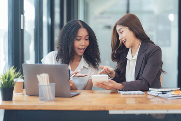 Obraz premium Joyful Collaboration: Two diverse businesswomen working together on a project, using laptop and calculator, showcasing teamwork and success in a modern office setting. 
