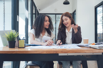 Collaborative Design Session: Two young, diverse female colleagues engage in a lively discussion over blueprints, showcasing teamwork and creative collaboration in a modern office setting. 