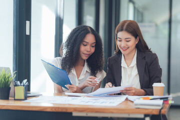 Collaborative Teamwork: Two diverse businesswomen, one Black and one Asian, engage in a focused discussion over documents and blueprints, showcasing their partnership and shared vision.  
