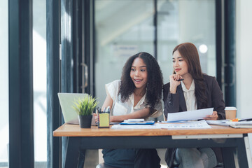 Obraz premium Collaborative Brainstorming: A diverse team of two young businesswomen strategize and discuss ideas, leaning in over a laptop and documents.