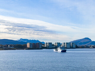Passenger ferry approaching the modern city coastline, with snow-capped mountains and cloudy sky in the background