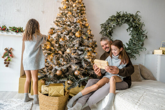 Festive family decorating Christmas tree. Father and two daughters share a joyful moment opening presents. Warm holiday atmosphere.