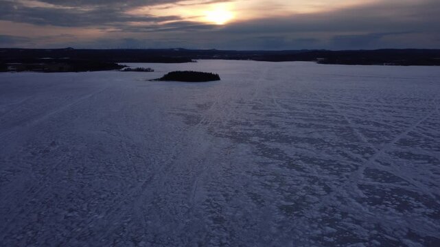 Dramatic aerial shot of a frozen lake, slowly tilting up revealing the setting sun behind the clouds in the distance. Snowmobile tracks making patterns in the ice.