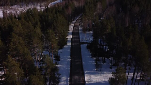 Aerial shot of a lone road surrounded by pine trees slowly tilting up revealing a calm, beautiful winter landscape