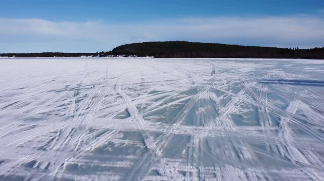 Camera slowly tilting up from the frozen lake covered in snowmobile tracks, to reveal a blue sky and small mountain in the distance