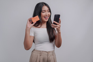 Pretty young Asian woman holding a credit card while looking at the smartphone she holds.