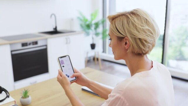 Caucasian woman in her 30s using smartphone to shop for clothing online in bright, modern kitchen. She sits at wooden table, engaged in browsing delivery website.