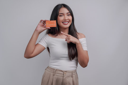 Cheerful young Asian woman pointing to the credit card she holds, isolated by white background.