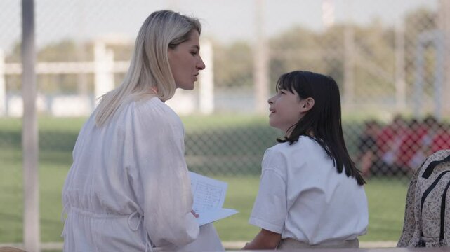 A caring mother helps her daughter with schoolwork on a sports field, fostering a strong bond while supporting her education. After school activities and family time.