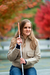 A visually impaired woman with a white cane sitting on a bench in a park on an autumn day