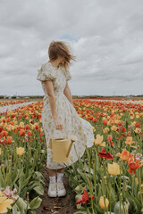 A girl in a yellow dress and apron with a water can in a red and green tulip flower field on a gloomy rainy cloudy sky background happily running around. 