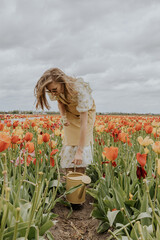 A girl in a yellow dress and apron with a water can in a red and green tulip flower field on a gloomy rainy cloudy sky background happily running around. 