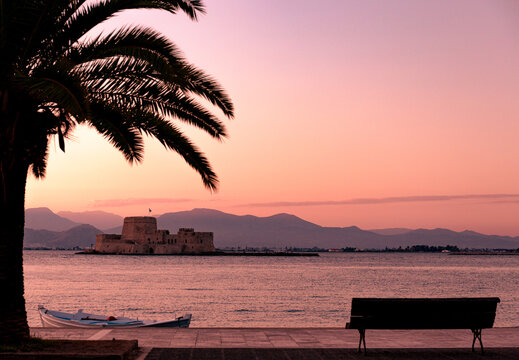 Postcard framed photo of beautiful and romantic walkway in Nafplio with Bourtzi fortress in the background and sun setting in golden and purple tones over the mountains.