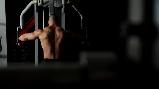A bodybuilder uses the fly simulator to train his pectoral muscles. Upper body workout in a dark gym, rear view. A handsome bodybuilder trains in the gym, shadows on his back draw every muscle.