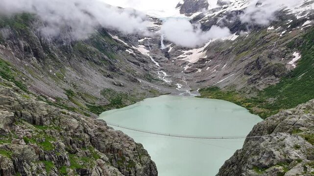 Turquoise Triftsee lake and suspension pedestrian bridge surrounded by swiss alps and clouds and glacial lake, Triftbr&uuml;cke Triftgletscher, Bern, Switzerland, pulling out aerial footage