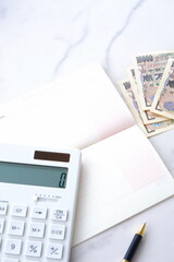 テーブルの上に開いた状態で置かれた銀行の預金通帳と、1万円札、電卓

