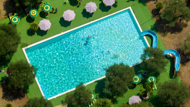 Aerial view of a rectangular pool in the garden of a resort. There is a slide for children and umbrellas for parents.