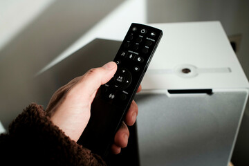 Close-up of a hand holding a black remote control, pointing towards a white speaker with a circular feature.