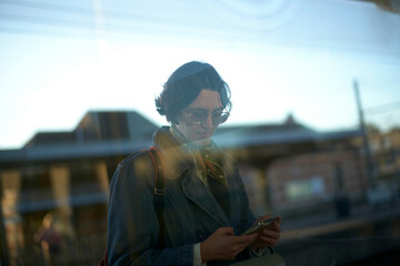 Young woman using a smartphone behind a window, with reflections of the urban environment overlaying their image.