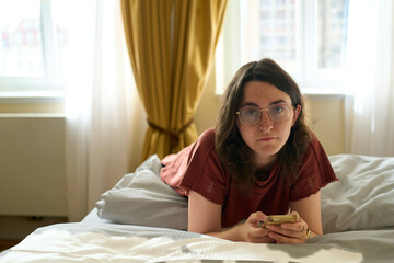 Young woman lying on a hotel bed while using her smartphone, with curtains and a window in the background, Dresden, Germany