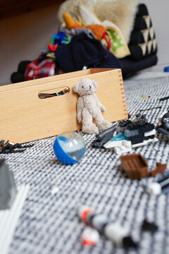A teddy bear leans against a wooden toy box amidst scattered toys on a patterned carpet, Germany