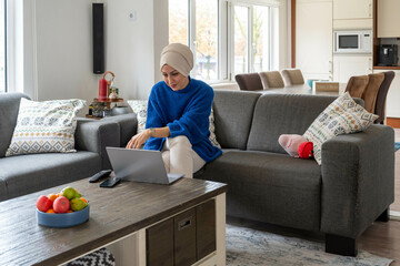 A woman in a blue sweater and hijab is sitting on a gray sofa, using a laptop with a bowl of fruit on the table in a modern living room.