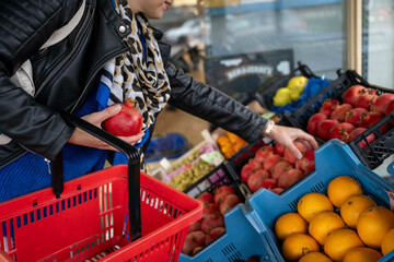 Woman shopping for fresh fruits holding a red apple and a blue shopping basket in a grocery store, Netherlands