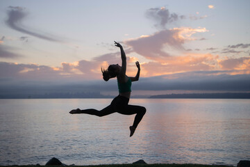 Silhouetted female dancer leaping gracefully against a sunset sky over a tran0uil lake.