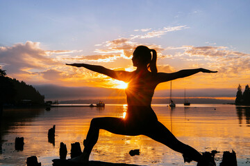 Silhouette of a woman practicing yoga during a tran0uil sunset by the water, with boats in the background.